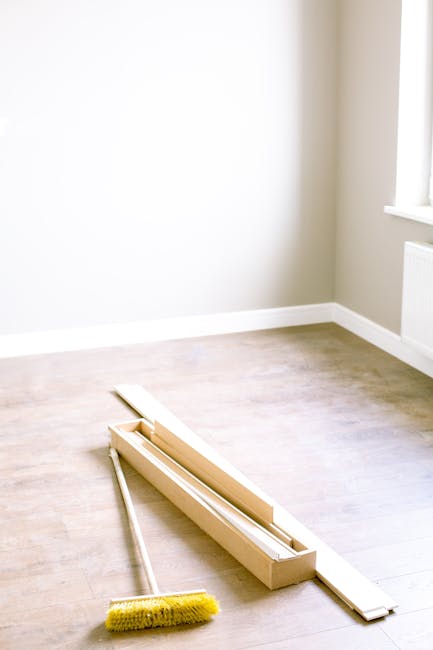 A clean, empty room with light-colored wooden flooring and beige walls illuminated by natural light coming through a window. On the floor, there is a wooden window blind installation kit, including a 