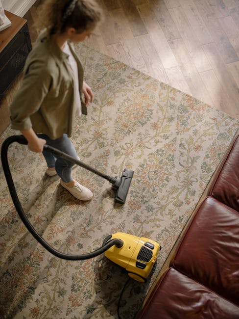 A professional cleaner from Cleaner Tooting, dressed in full protective gear including a white suit, gloves, face mask, and shoe covers, is performing deep cleaning on a beige, plush carpet in a brigh